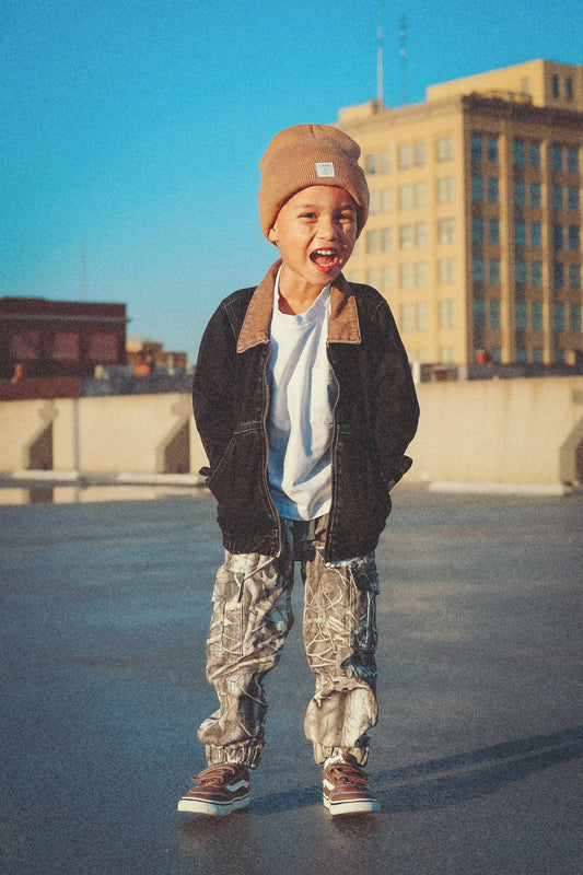 Child wearing a brown beanie, black jacket, white shirt, camouflage pants, and brown shoes.