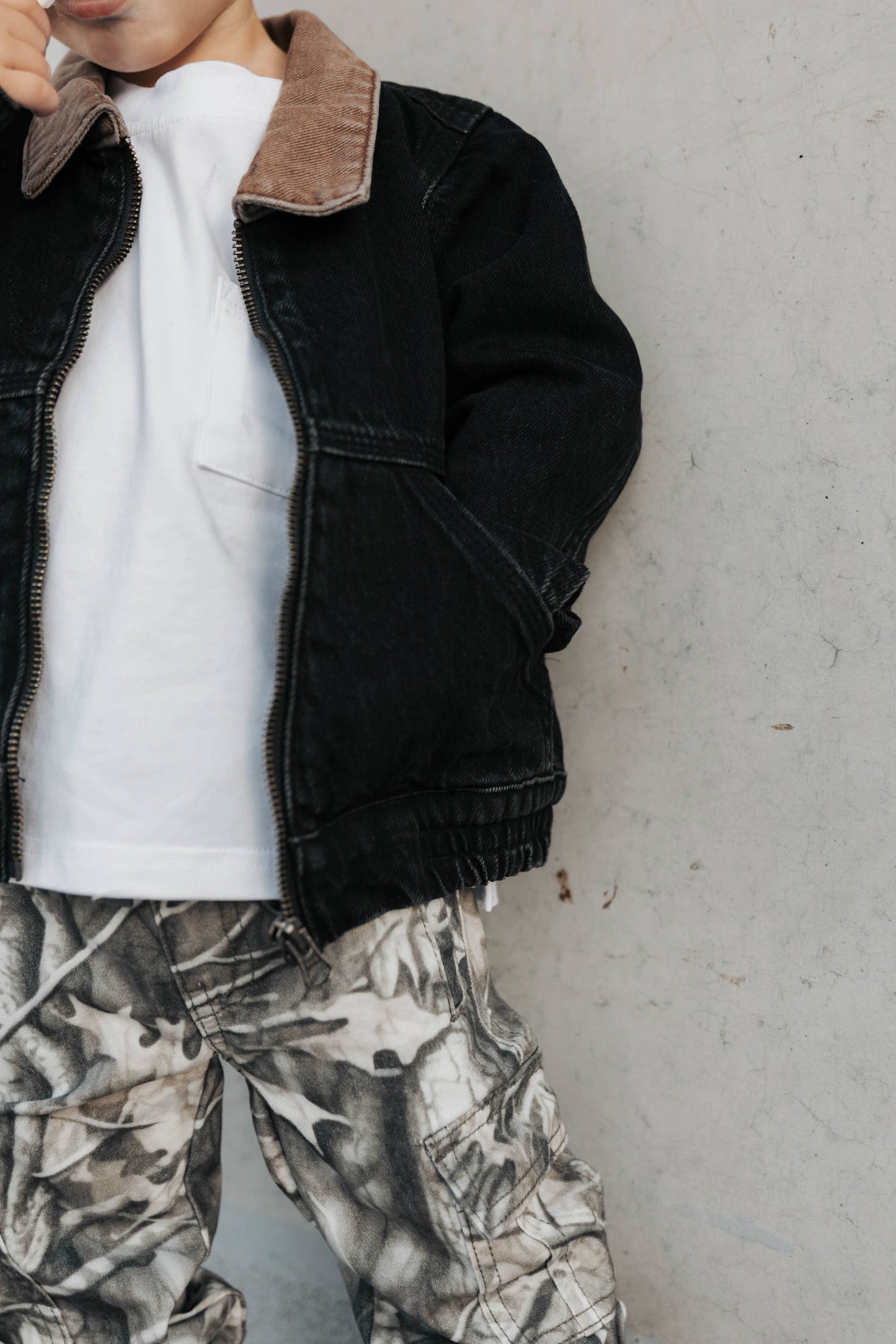 Kid wearing a black denim utility jacket, white shirt, and camouflage pants.