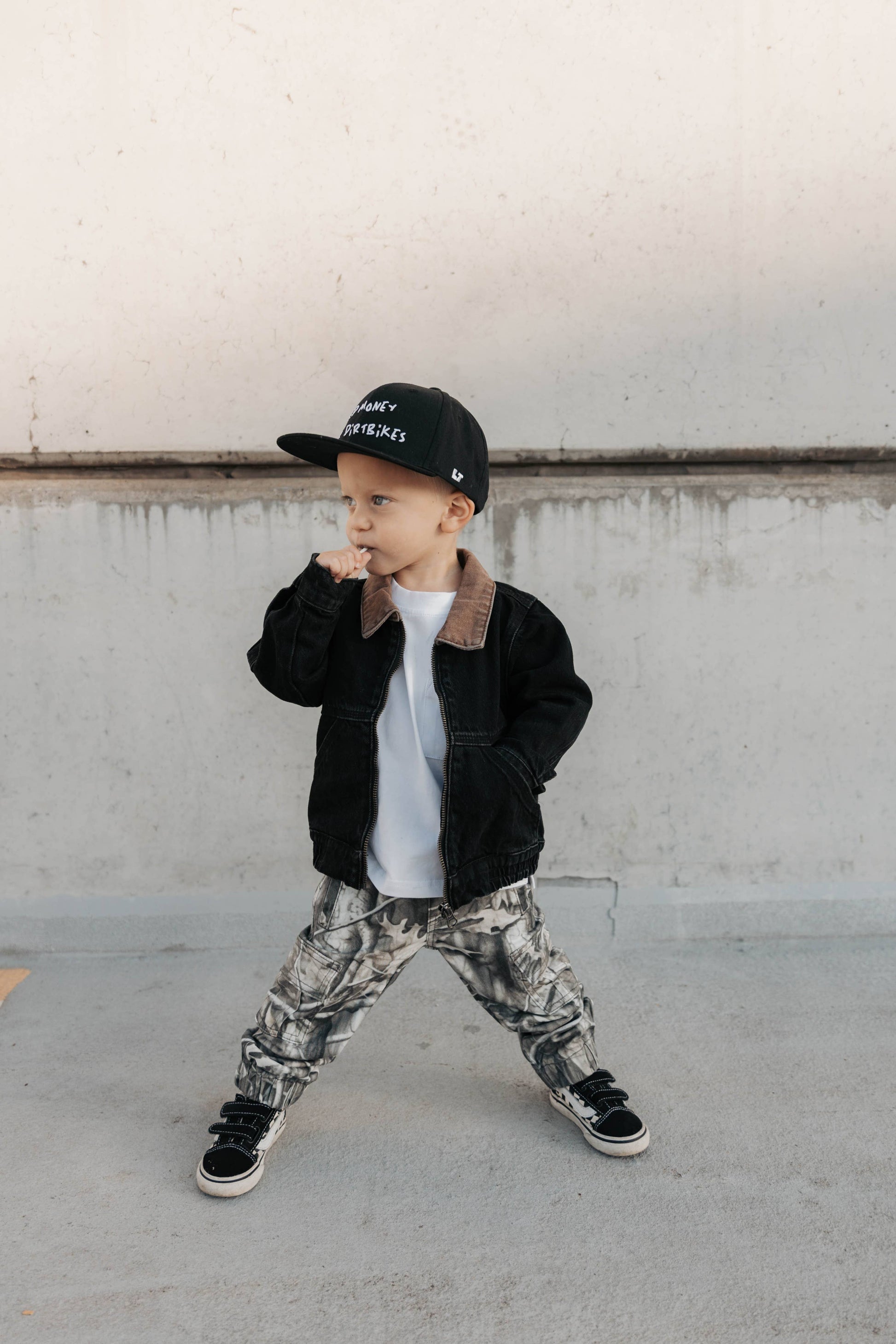 Child wearing a black denim utility jacket, white shirt, camouflage pants.